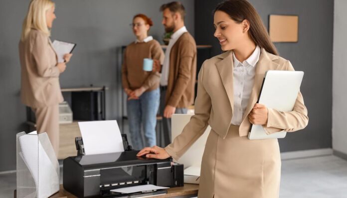 Young businesswoman with laptop using modern printer in office